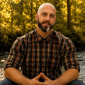 Man with a beard in a plaid shirt sitting outdoors with a forest background.
