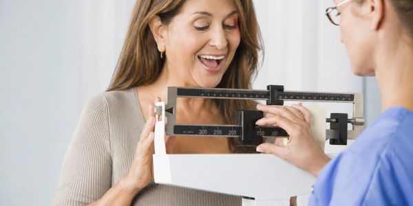 A happy woman being weighed on a mechanical scale by a healthcare professional.
