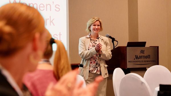 A woman in a beige suit claps while standing near a podium at a Marriott hotel event.