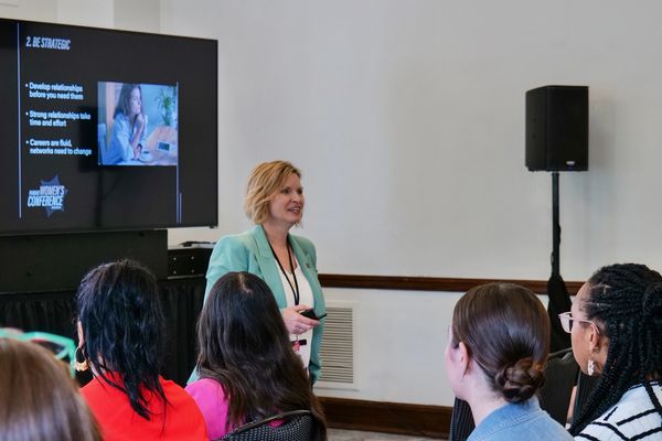 A woman in a teal blazer leads a women's conference presentation to an attentive audience.