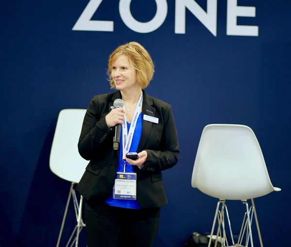 A woman holding a microphone and remote, standing in front of chairs and a blue background.