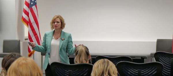 Woman in teal blazer giving a presentation to an audience with an American flag in the background.