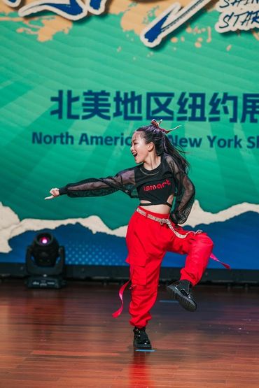 A young dancer performs energetically on stage with vibrant red pants and a black top.