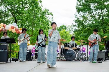 A young band performs outdoors with instruments and microphone amid green trees.