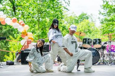 Three young dancers in white outfits pose on an outdoor stage with musical instruments.