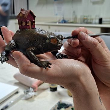 Artist painting a detailed frog sculpture with a tiny house on its back.