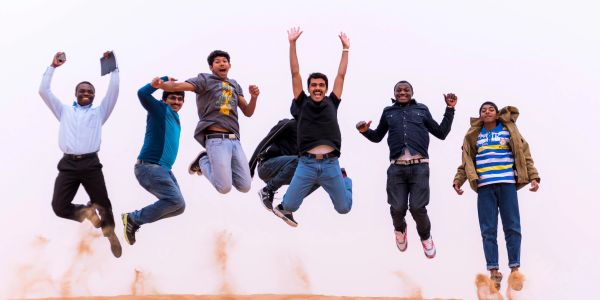 Seven men joyfully jump mid-air with dust rising beneath them against a clear sky.