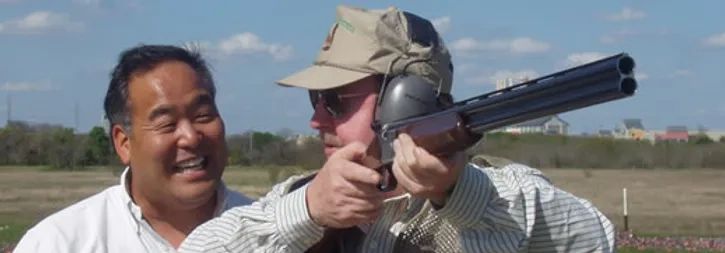 Two men outdoors, one aiming a shotgun while the other smiles beside him.
