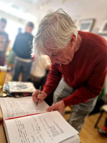 Older man in red sweater writing in a notebook at a busy indoor event.