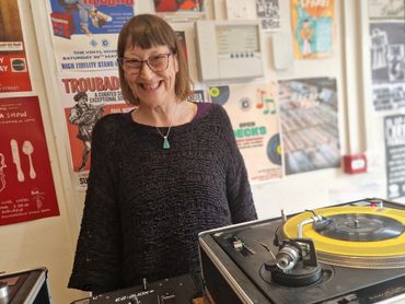 A smiling woman stands behind a DJ turntable in a room filled with vintage posters.