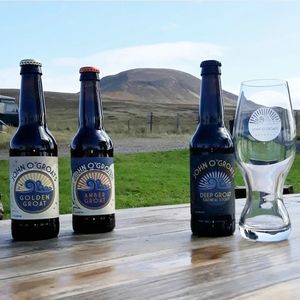 Three John O'Groats beer bottles and a branded glass on a wooden table outdoors.