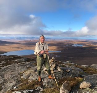 Man standing on rocky hilltop with walking stick against vast moorland and lake.