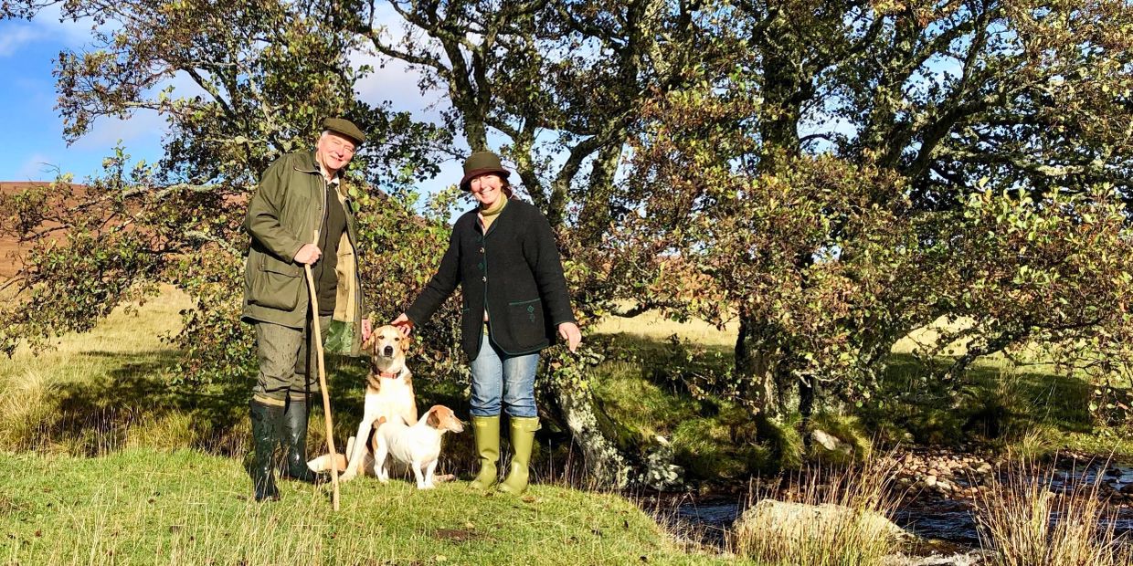 A smiling couple with two dogs standing outdoors near a tree and stream on a sunny day.