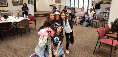 A group of our students, in a class, huddled together and smiling in their talk to the camera Tees.