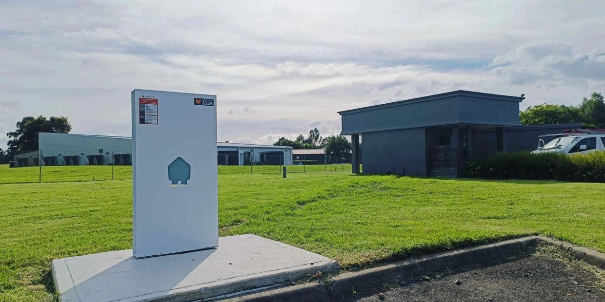 A white utility box stands on a concrete slab in a grassy area near buildings.