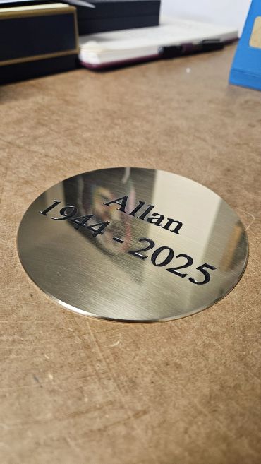Circular metallic plaque engraved with "Allan 1944 - 2025"