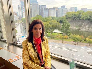 Woman in a gold jacket sitting by a window with a cityscape and greenery outside.
