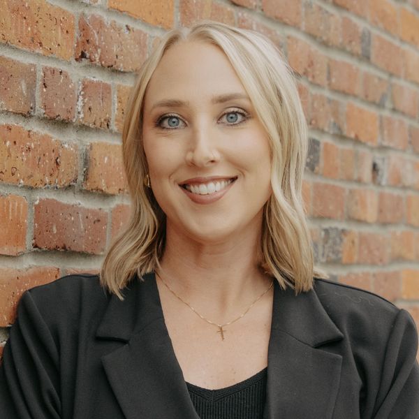 Smiling woman with blonde hair in black blazer against brick wall.