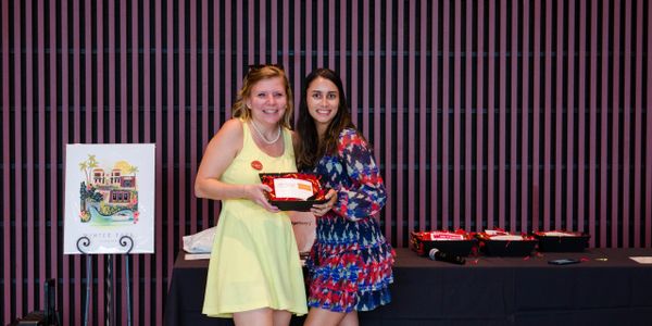 Two smiling women holding a red tray in front of a striped wall and table.