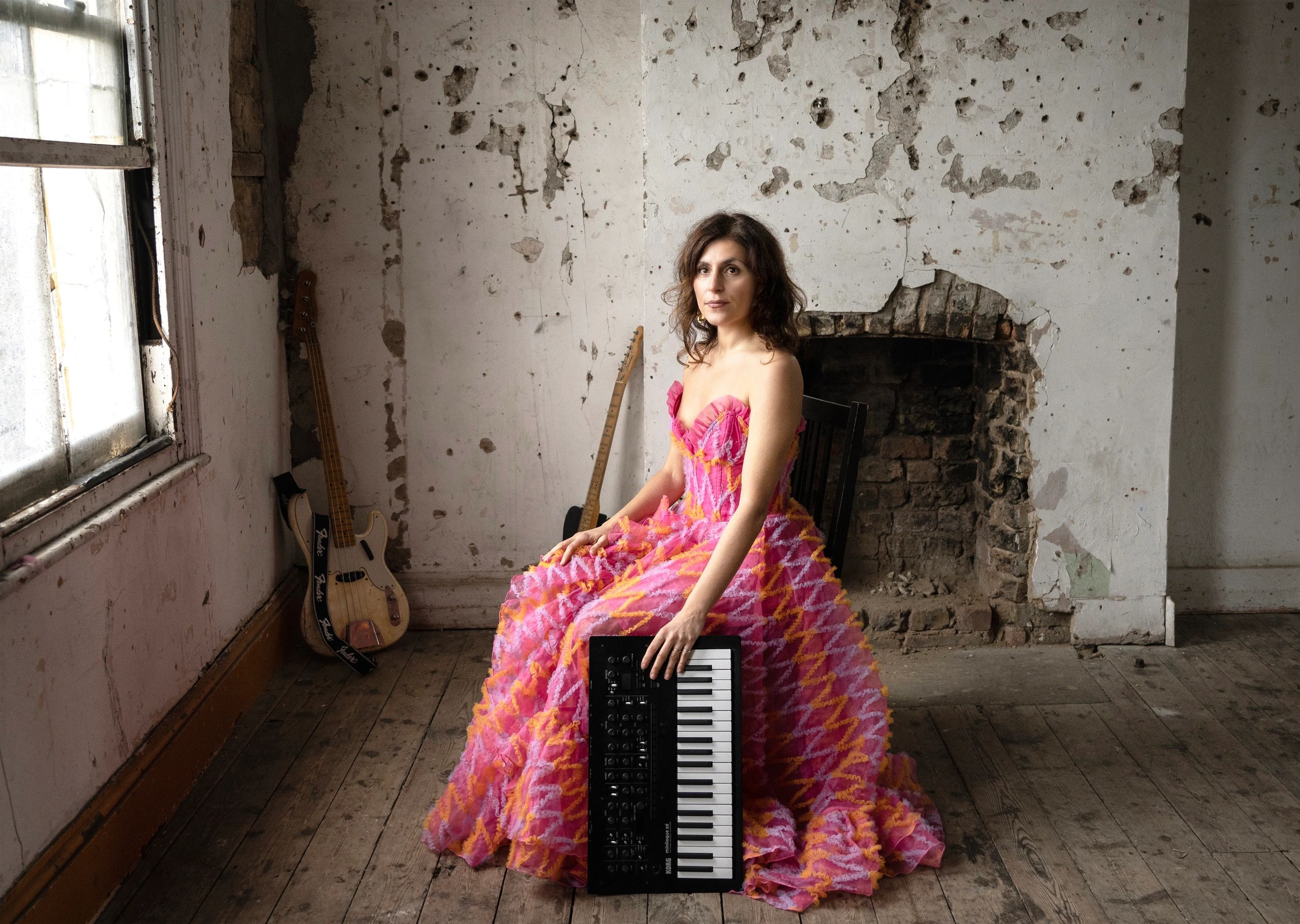 Woman in a vibrant dress holding a keyboard, sitting in a rustic room with musical instruments.