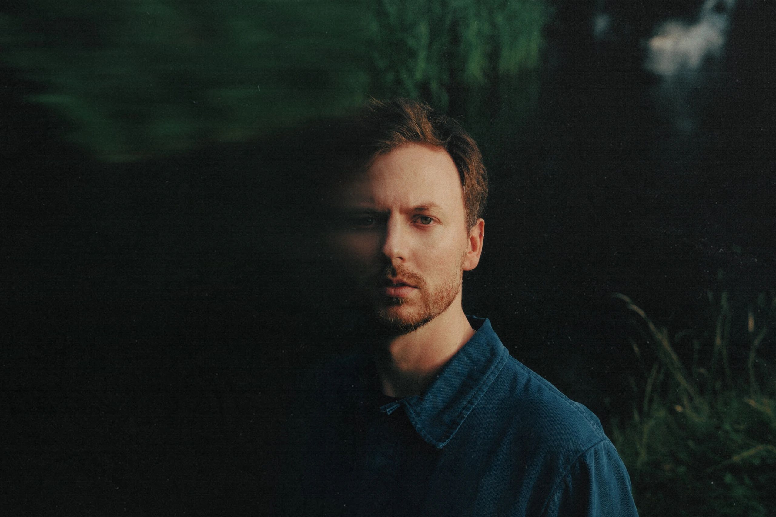 Portrait of a man partially obscured by shadow against a dark, natural background.