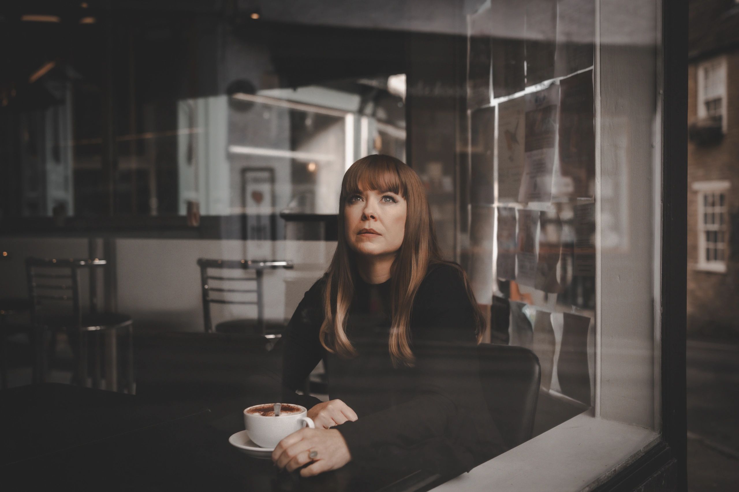 A woman with long hair looks thoughtfully out a cafe window, holding a cup of coffee.