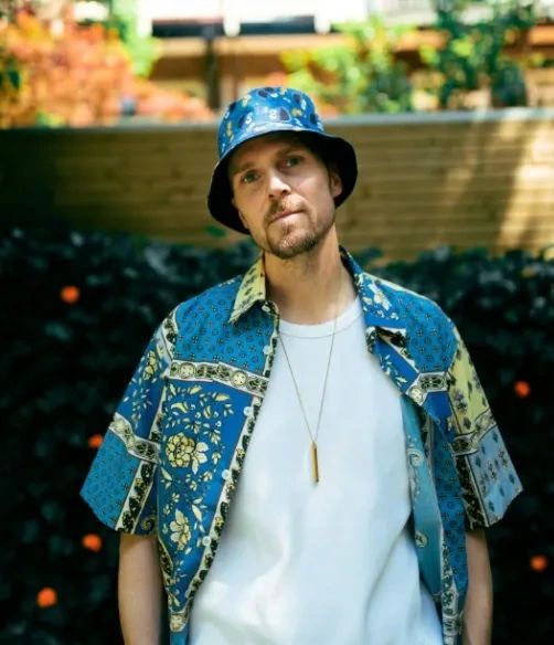 Man wearing a blue patterned shirt and bucket hat standing outdoors.