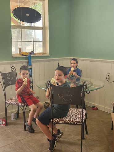 Three boys sitting around a glass table eating ice cream cones indoors.