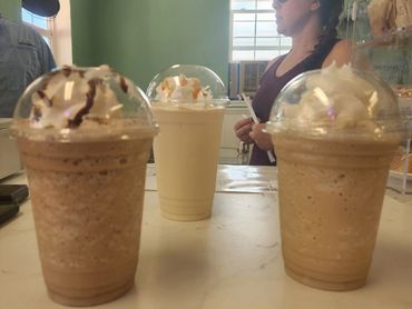 Three iced coffee drinks with whipped cream on a counter, a woman in sunglasses in background.