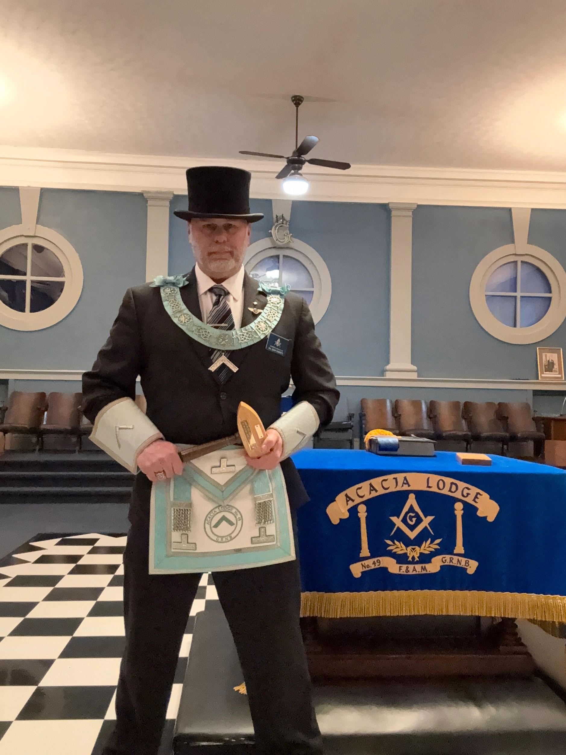 Man in formal Masonic regalia holding ceremonial tools in a lodge room.