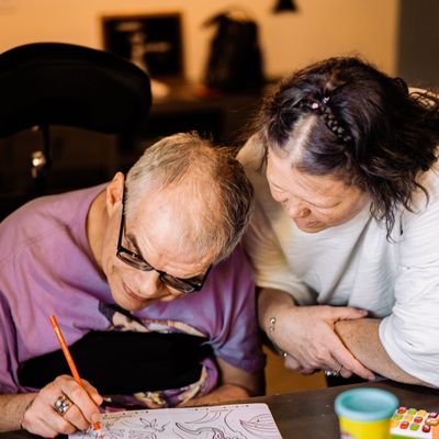 A support staff leaning in next to a person she supports watching him color with colored pencils.