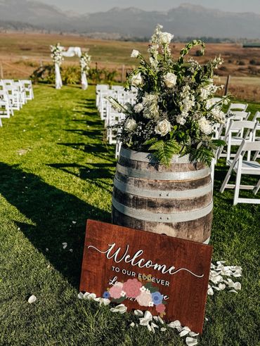 Rustic wedding setup with a floral arrangement on a barrel and a welcome sign on the grass.
