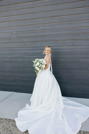 Bride in a flowing white wedding dress holding a bouquet, smiling against a dark wooden wall.