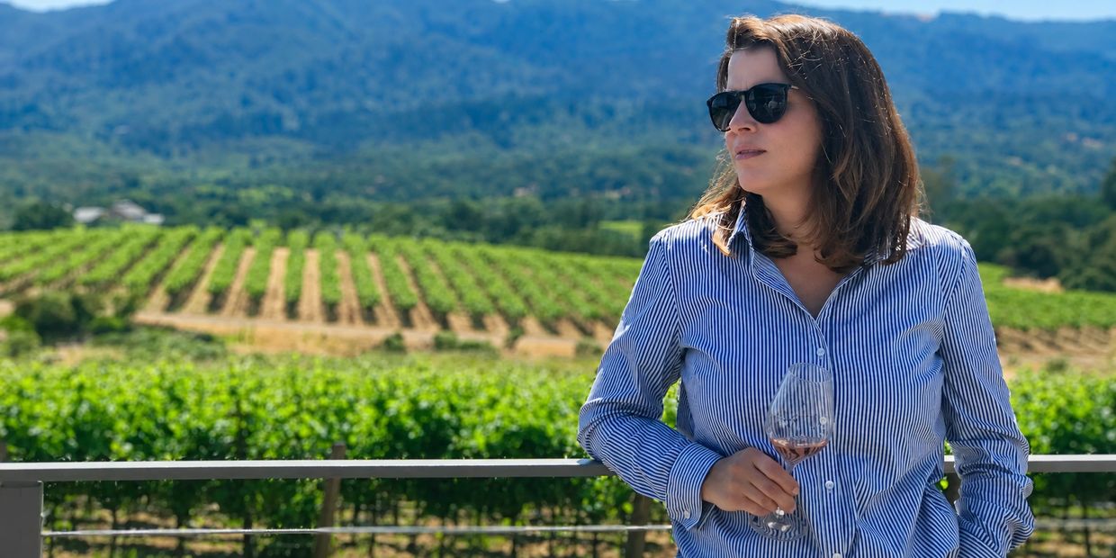 Woman in sunglasses enjoying wine at a vineyard with scenic mountain views.