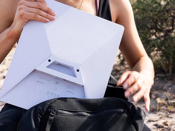 Person placing a white electronic device into a black backpack outdoors.