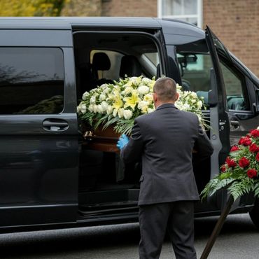 Man in suit loading a flower-adorned coffin into a black van.