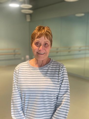 Smiling elderly woman in a striped shirt stands in a spacious dance studio.