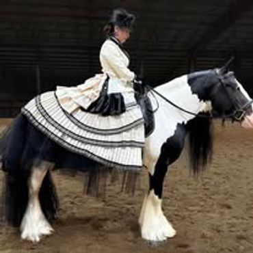 A woman in Victorian attire rides a black and white horse indoors.