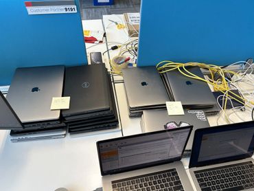Stacks of Apple and Dell laptops on a desk with notes and tangled cables.