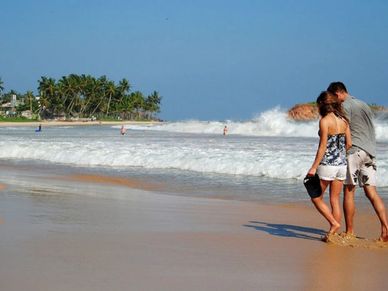 Couple walking barefoot on a sunny beach with waves crashing.