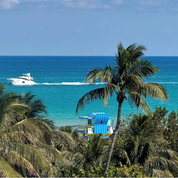 Palm trees frame a bright blue lifeguard tower by the turquoise ocean with a yacht in the distance.