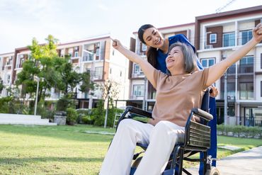 A caregiver assists a person in a wheelchair, both smiling outdoors in a well-maintained grassy area