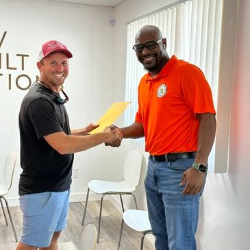 Two men shaking hands indoors, one holding an envelope.