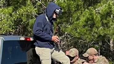 Military personnel in camouflage gear around pickup trucks in a wooded area.