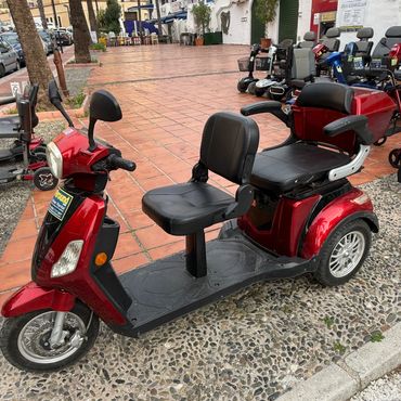 A red mobility scooter with two seats parked on a stone and tile sidewalk.