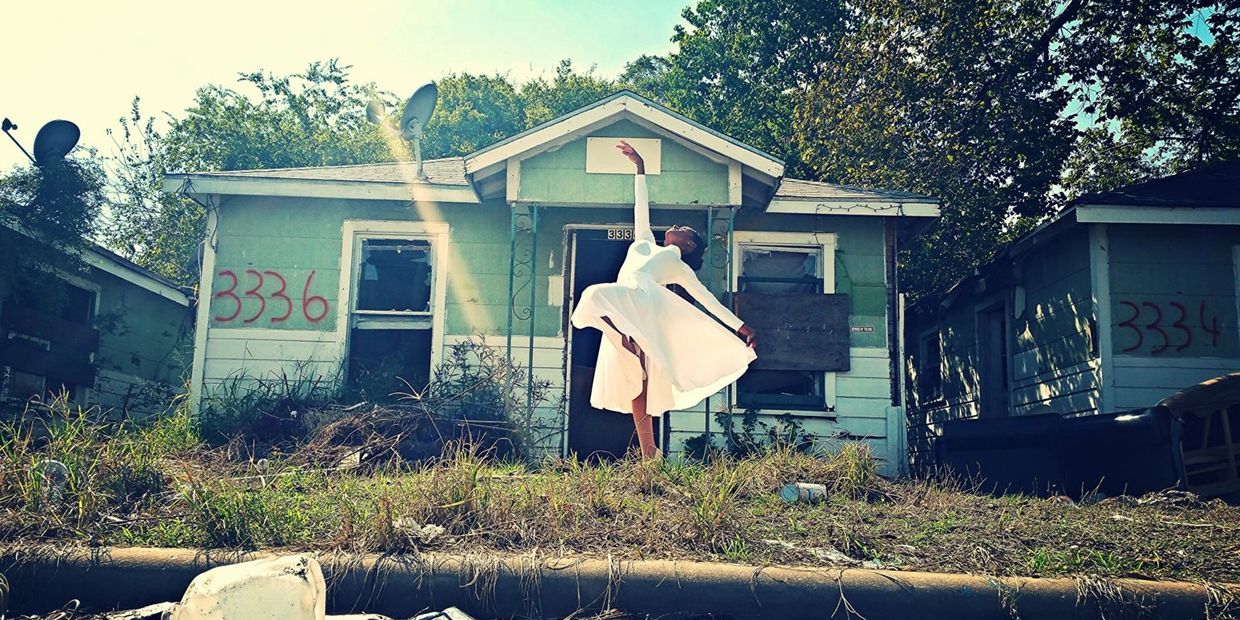 A dancer in white poses gracefully in front of a rundown house with broken windows.