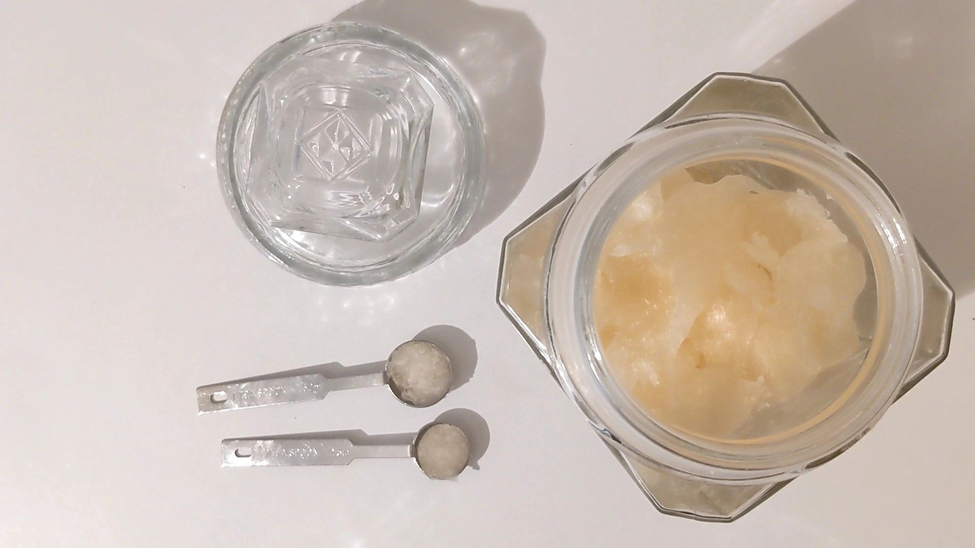 Glass jar with a creamy substance, two metal measuring spoons, and a glass on a white surface.