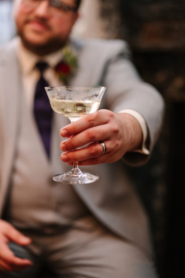 Groom in gray suit holding a coupe glass with a sparkling drink. Wedding ring visible.