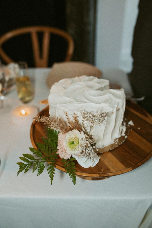 Small, white wedding cake decorated with flowers and greenery on a wooden platter on a white table