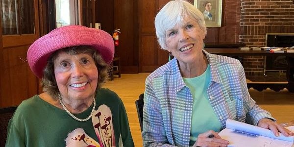 Two elderly women smiling indoors, one wearing a pink hat.